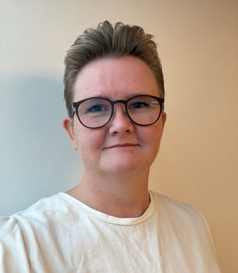Person med briller og kort hår i hvid bluse.